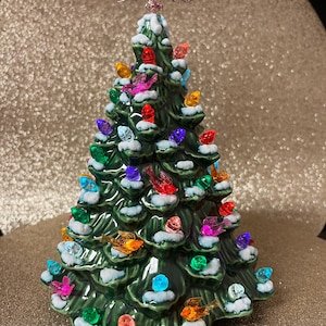 May include: A ceramic Christmas tree in green, decorated with multi-coloured lights and bird ornaments. The tree is topped with a glittery star. The branches have a snow-like effect. The tree is a festive decoration.