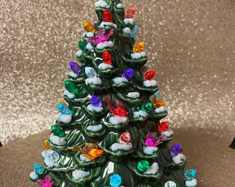 May include: A ceramic Christmas tree with a green glaze, adorned with colorful lights and small bird ornaments. The tree is topped with a glittery star. The tree has a snow-like effect on the branches.