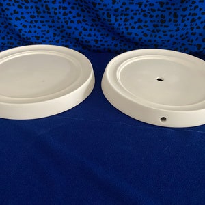 May include: Two white ceramic round cake stands with a hole in the centre. The stands are on a blue background.