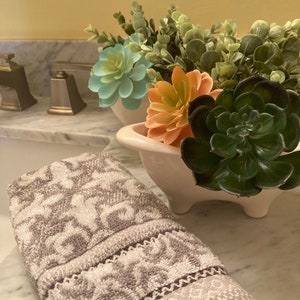 May include: A white ceramic planter with a decorative, scalloped edge, filled with a variety of green succulents and artificial flowers. The planter is sitting on a marble countertop.