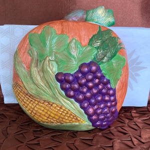 May include: Ceramic pumpkin-shaped napkin holder with a 3D design of grapes, corn, and leaves. The pumpkin is orange, the grapes are purple, the corn is yellow, and the leaves are green.