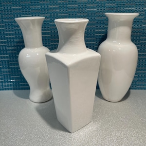 May include: Three white ceramic vases in different shapes. The vase on the left has a curved form. The central vase is geometric and angular. The vase on the right has a rounded shape. All vases have a glossy finish, suitable for floral arrangements.