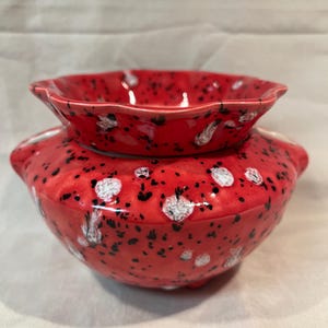 May include: A two-piece ceramic bowl set. The base bowl is a vibrant red with black and white speckled accents. The top bowl is smaller, with a scalloped edge, and also red with speckled details. The set is likely for serving or display.