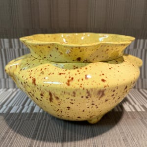 May include: A yellow ceramic bowl with a speckled brown pattern, featuring a wide, scalloped rim and a rounded body. The bowl is supported by small, integrated feet and has a glossy finish, showcasing a distinct two-piece design structure.