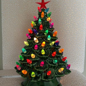 May include: A ceramic Christmas tree in a deep green colour, adorned with colourful, glowing lights. A red star sits atop the tree. The tree is lit with a variety of coloured lights, including red, yellow, green, and blue.
