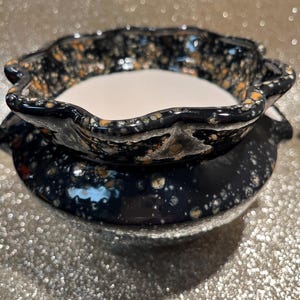 May include: A two-tiered ceramic candle holder with a scalloped rim. The holder is navy blue with speckled orange and white accents. The interior of the top tier is white, and the base is a solid navy blue.