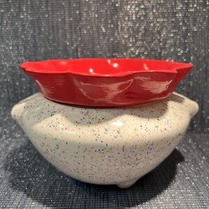May include: A red ceramic bowl with a scalloped edge sits on a speckled white ceramic pot. The pot has a rounded shape with small, decorative feet. The background is a textured, dark blue surface.
