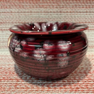 May include: A handcrafted, round ceramic bowl with a glossy, deep red glaze. The bowl features a flared rim and a textured exterior with white and black accents, creating a unique, artistic appearance. The bowl is suitable for decorative or functional use.