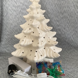 Puede incluir: Un árbol de Navidad de cerámica blanca con agujeros para luces. El árbol está rodeado por un kit de luces, una bolsa de luces de colores y un adorno de estrella verde. El árbol no está pintado y está listo para personalizar.