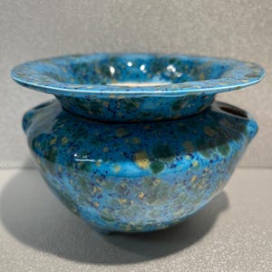 May include: A turquoise and blue ceramic bowl with a wide rim and rounded base. The glaze features speckled yellow and green accents. The rim has a slightly raised edge. The bowl is likely handmade.