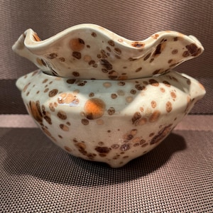 May include: A two-piece ceramic bowl set. The bowls are off-white with brown and orange speckled patterns. The top bowl has a ruffled edge, and the bottom bowl has a rounded shape.