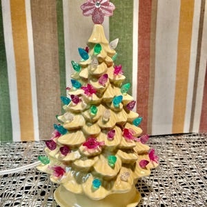 May include: A cream-coloured ceramic Christmas tree, decorated with multi-coloured lights and a pink star topper. The lights are in shades of pink, green, blue, and clear. The tree is plugged in with a white cord, and is a festive decoration.