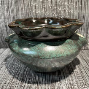 Peut inclure: Ensemble de poterie en céramique en deux parties. La pièce du bas est un grand bol vert-turquoise avec une surface texturée. La pièce du haut est un petit bol brun foncé avec un bord festonné. Les deux pièces ont une finition brillante.