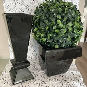 May include: Two black ceramic planters, one tall and narrow, the other shorter and wider. The shorter planter holds a small green artificial topiary.