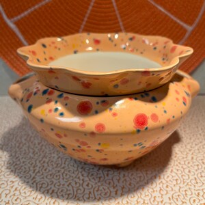 May include: Two-piece ceramic serving bowl set. The bowls are peach-coloured with colourful speckled designs. The top bowl has a scalloped edge. The bottom bowl has two handles.