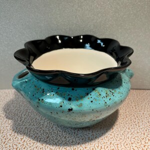 May include: A two-piece ceramic decorative bowl set. The bottom bowl is turquoise with gold speckles and handles. The top bowl is black with a scalloped edge and a white interior. The set is designed for decorative purposes.