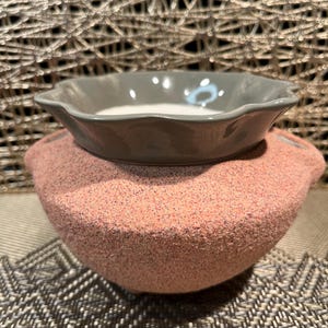 May include: A two-piece ceramic wax warmer. The top dish is grey with a scalloped edge, and the base is a textured pink. The warmer is designed to hold wax melts for fragrance.
