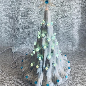 May include: A white ceramic tabletop Christmas tree with a textured, snowy finish. The tree is illuminated by small, glowing light bulbs and decorated with bright blue circular accents. A clear, glittery snowflake topper with a blue centre sits at the top.