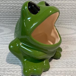 Peut inclure: Une figurine de grenouille en céramique vert brillant avec une large bouche ouverte. La grenouille a de grands yeux noirs lisses et un intérieur de couleur crème clair. La sculpture est représentée en position assise.