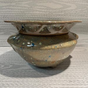 May include: A handmade ceramic pot with a matching lid. The pot is a rounded shape with a wide base and a slightly flared rim. The lid is a shallow dish with a wide, flat edge. The pot and lid are glazed in shades of brown, tan, and blue.