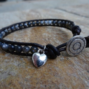 May include: A brown leather bracelet with a silver heart charm and a silver circle charm. The bracelet is made with dark gray beads.