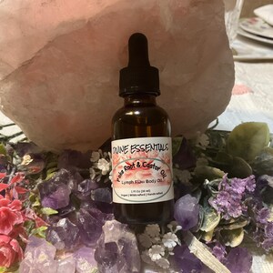 May include: A brown glass bottle with a dropper top filled with a clear liquid. The bottle label reads "Divine Essentials Poke Root & Castor Oil Lymph Detox Body Oil 1 fl oz (30 ml) Vegan, Wildcrafted, Handcrafted". The bottle is surrounded by purple and pink crystals and flowers.