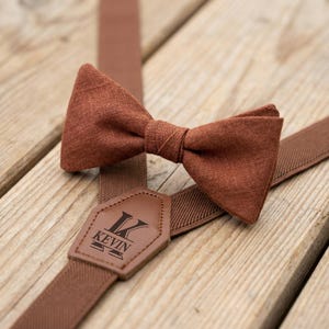 May include: A brown bow tie and suspenders set. The bow tie is made of textured fabric. The suspenders have a leather patch with the name "KEVIN" and a stylized "K" initial. The set is displayed on a wooden surface.