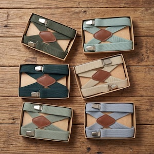 Könnte beinhalten: Sechs Hosenträgerpaare in verschiedenen Farben, darunter Grün, Türkis, Beige und Blau, werden in einzelnen Pappschachteln präsentiert. Jedes Hosenträger-Set verfügt über einen rautenförmigen Aufnäher aus braunem Leder und silberfarbene Metallverschlüsse.