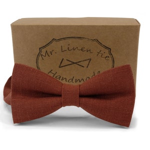 Op de afbeelding: Een roestkleurige linnen vlinderdas met een getextureerde uitstraling, gepresenteerd voor een bruine kartonnen doos. De doos heeft een gestempeld logo met de tekst "Mr. Linen tie Handmade". Een klassiek accessoire.