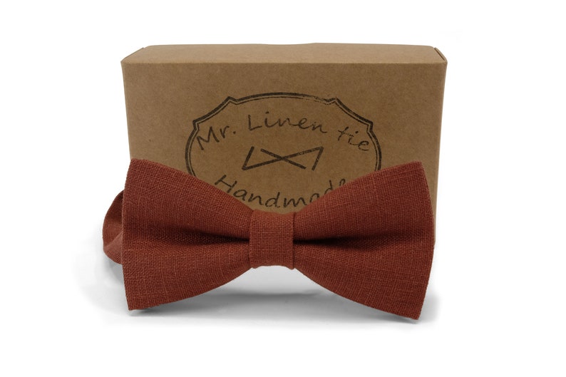 Op de afbeelding: Een roestkleurige linnen vlinderdas met een getextureerde uitstraling, gepresenteerd voor een bruine kartonnen doos. De doos heeft een gestempeld logo met de tekst "Mr. Linen tie Handmade". Een klassiek accessoire.