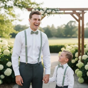 Può includere: Un uomo e un ragazzo, entrambi con camicie bianche, papillon e bretelle, sono in piedi all'aperto. L'uomo indossa una camicia bianca, pantaloni grigio scuro e bretelle e papillon verde oliva. Il ragazzo indossa un abito simile. Si tengono per mano e sorridono.