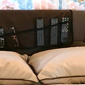 May include: A black fabric organizer with five pockets, holding four black remote controls and a pair of glasses. The organizer is attached to the headboard of a bed with two beige pillows.
