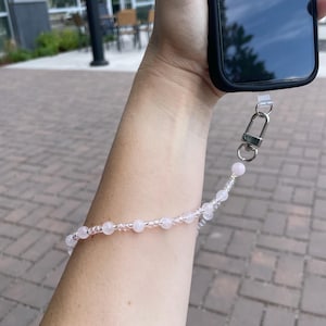 May include: A beaded phone wrist strap featuring alternating light pink round beads and small clear faceted beads. The strap is attached to a black smartphone case via a silver-toned metal lobster clasp and a clear plastic connector loop.