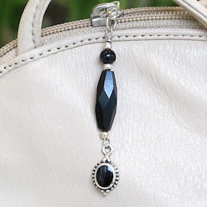 May include: A black onyx charm with a silver chain and a silver oval pendant with a black onyx stone. The charm is attached to a light beige purse.