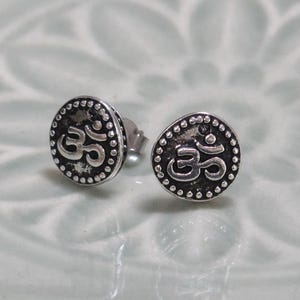 May include: A pair of round silver stud earrings featuring a raised Om symbol in the center. The circular design is bordered by a series of small, raised silver dots, and the metal has a dark, oxidized finish in the recessed areas.