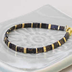 Puede incluir: Un brazalete negro y dorado con un patrón geométrico. El brazalete está hecho de pequeñas baldosas cuadradas con un borde dorado.