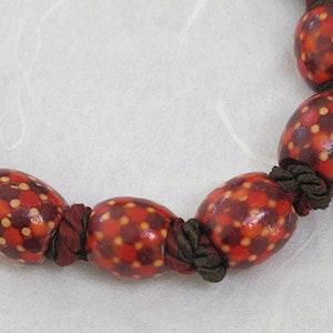 May include: A close-up of a necklace featuring large, oval-shaped beads. The beads are primarily red with a pattern of small yellow and orange dots. The beads are strung together with dark brown cord.