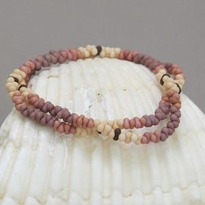May include: A multi-colored beaded bracelet with shades of pink, brown, and cream. The bracelet is made of small, round beads and is displayed on a white seashell.