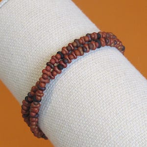 May include: A beaded bracelet with a double strand design. The beads are a mix of brown and black, creating a textured appearance. The bracelet is displayed on a white fabric background against an orange backdrop.