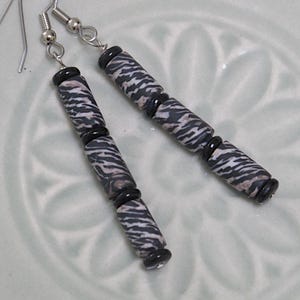 May include: A pair of dangle earrings featuring long, cylindrical beads with a black, white, and grey zebra-like striped pattern, separated by small, smooth, round black spacer beads, suspended from silver-toned metal earring hooks.