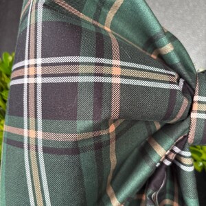 May include: Close-up of a green plaid bow tie. The fabric features a classic tartan pattern with lines of white, peach, and black. The bow is knotted and has a textured appearance, suggesting a soft material.