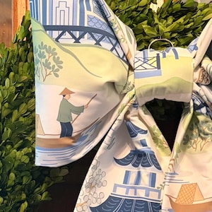 Puede incluir: Un lazo de tela grande con un estampado estilo toile en azul, verde y blanco. El patrón muestra escenas de pagodas, puentes y una persona con sombrero cónico remando en un bote sobre el agua. El lazo está sujeto con un nudo central y un anillo.