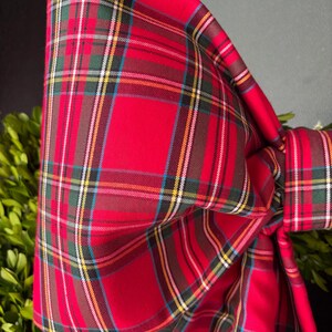 May include: A large decorative bow made of fabric featuring a red, green, yellow, and white plaid tartan pattern. The bow is tied with a central knot and has two long, flowing tails extending downwards, showcasing the repeating geometric grid design.