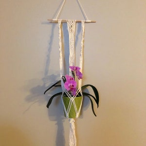 May include: A white macrame plant hanger with a wooden dowel and a green pot with a pink orchid plant.