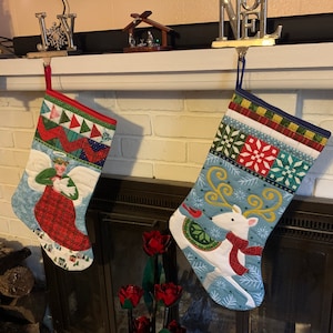 May include: Two Christmas stockings hanging on a white mantel above a fireplace. The stocking on the left features a blue background with an angel holding a dove. The stocking on the right features a blue background with a white reindeer wearing a red scarf.