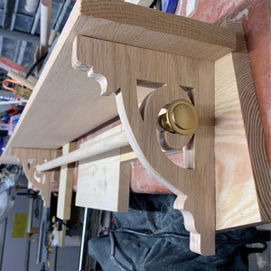 May include: A wooden shelf with decorative scrollwork brackets and a brass knob. The shelf is mounted to a wall and has a wooden dowel rod attached to the underside.