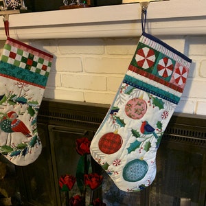 May include: Two Christmas stockings hanging on a white mantel above a fireplace. The stocking on the left is white with a red bird and green holly. The stocking on the right is white with red and pink ornaments and a blue bird.