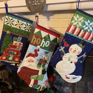 May include: Three Christmas stockings hanging on a white brick fireplace mantel. The stocking on the left has a red truck with presents on it. The middle stocking has a Santa Claus with presents and the text "ho ho ho". The stocking on the right has a snowman with a red bird and green garland.
