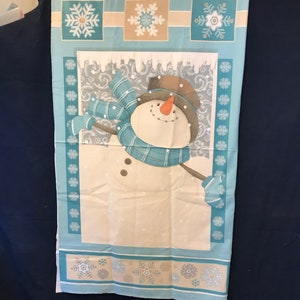 May include: A rectangular fabric panel featuring a central snowman illustration with a blue plaid scarf, brown hat, and orange carrot nose. The background includes white snowflakes on a light blue field, with decorative borders of snowflakes.