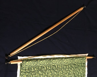 May include: A wooden dowel rod with black metal end caps and a gold cord attached. The rod is hanging from the ceiling and has a green and white patterned fabric attached to it.